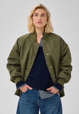 Femme aux cheveux blonds courts portant une veste bomber vert olive, un pull bleu marine, une chemise à carreaux et un jean bleu, les mains dans les poches.