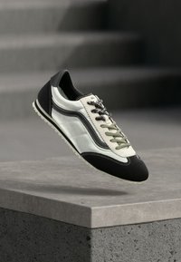 Baskets blanches et noires avec des lacets bicolores, flottant au-dessus d'un rebord en béton avec des escaliers flous en arrière-plan.