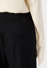 Černé kalhoty s hladkým povrchem, s jednou zadní kapsou a štítkem. V kombinaci se světle béžovou, lehce průhlednou blůzou.