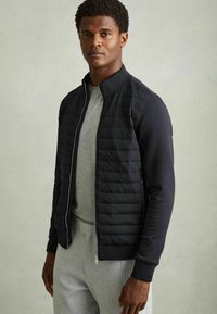 Jachetă puf din negru cu guler înalt și manșete elastice, cu față matlasată, asortată cu un hanorac gri și pantaloni gri deschis.