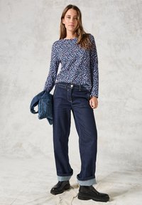 Langärmliges Hemd mit multicolorem abstraktem Muster, kombiniert mit lockeren dunklen Jeans und schwarzen Stiefeletten, während eine Denimjacke gehalten wird.