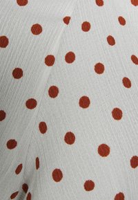 Tecido de algodão em branco com uma superfície texturizada, adornado com bolinhas de cor ferrugem de tamanhos variados, dispersas.