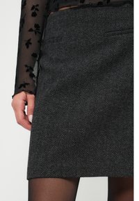 Saia em espinha de peixe carvão com corte reto, apresenta dois bolsos frontais e uma textura suave, combinada com uma blusa de mangas longas florais e translúcidas.