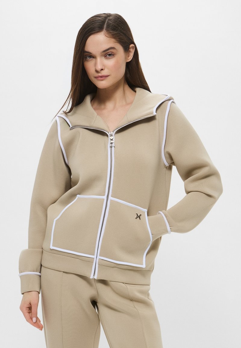 Felpa beige con zip e cappuccio, piping bianco, due tasche frontali e un piccolo logo vicino alla tasca. Presenta una texture liscia.