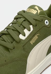 Κοντινό πλάνο πράσινου σουέντ αθλητικού παπουτσιού Puma με μπεζ ρίγα, πράσινα κορδόνια που ταιριάζουν και χρυσό λογότυπο Puma στη γλώσσα και στο πλάι.