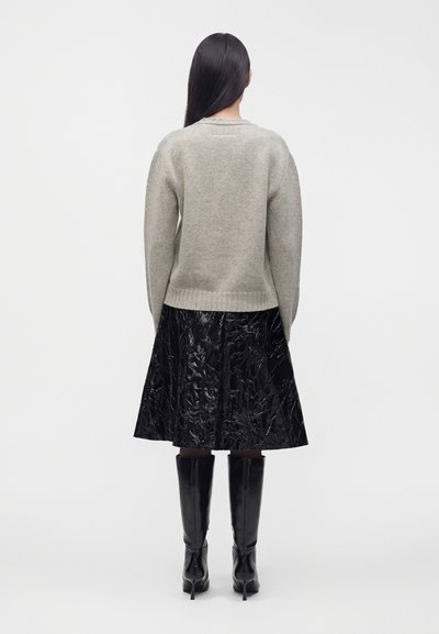 Femme aux longs cheveux noirs, vue de dos, portant un pull gris, une jupe noire brillante longueur genou et des bottes noires à talons hauts.