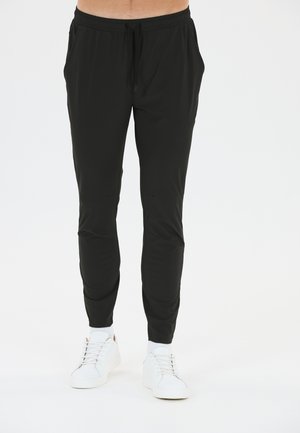 Mann trägt schwarze Slim-Fit-Jogginghosen mit Kordelzugbund und weißen Sneakern, steht vor weißem Hintergrund.