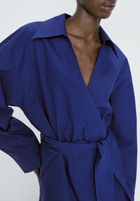 Lång, djupt blå omlottklänning av slätt tyg. Har en stor krage, vida ärmar och ett knytbälte i midjan. Rent design utan mönster.