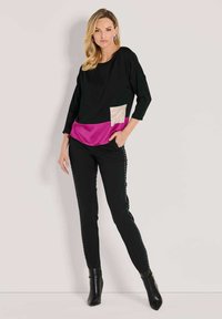 Frau in schwarzem Oberteil mit pinkem Saum und beigem Tascheneinsatz, schwarzen Hosen mit Seitennieten und schwarzen Stiefeletten, die vor einem einfarbigen Hintergrund steht.