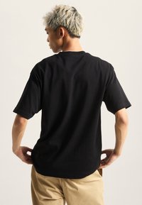 Černé bavlněné tričko s krátkým rukávem a volným střihem. Model má na sobě khaki kalhoty, které ukazují zadní pohled na tričko.