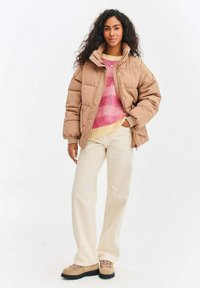 Hnedá prešívaná bunda s vysokým golierom, nosená cez ružový a smotanový pásikavý sveter, smotanové široké nohavice a béžové turistické topánky s čiernymi podrážkami.