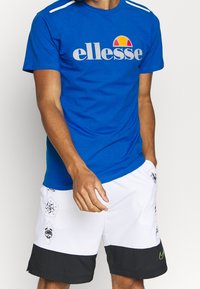 Camiseta de manga corta azul con el logo de "ellesse" en gris y naranja. Combinada con pantalones cortos blancos con dobladillo negro y acentos estampados.