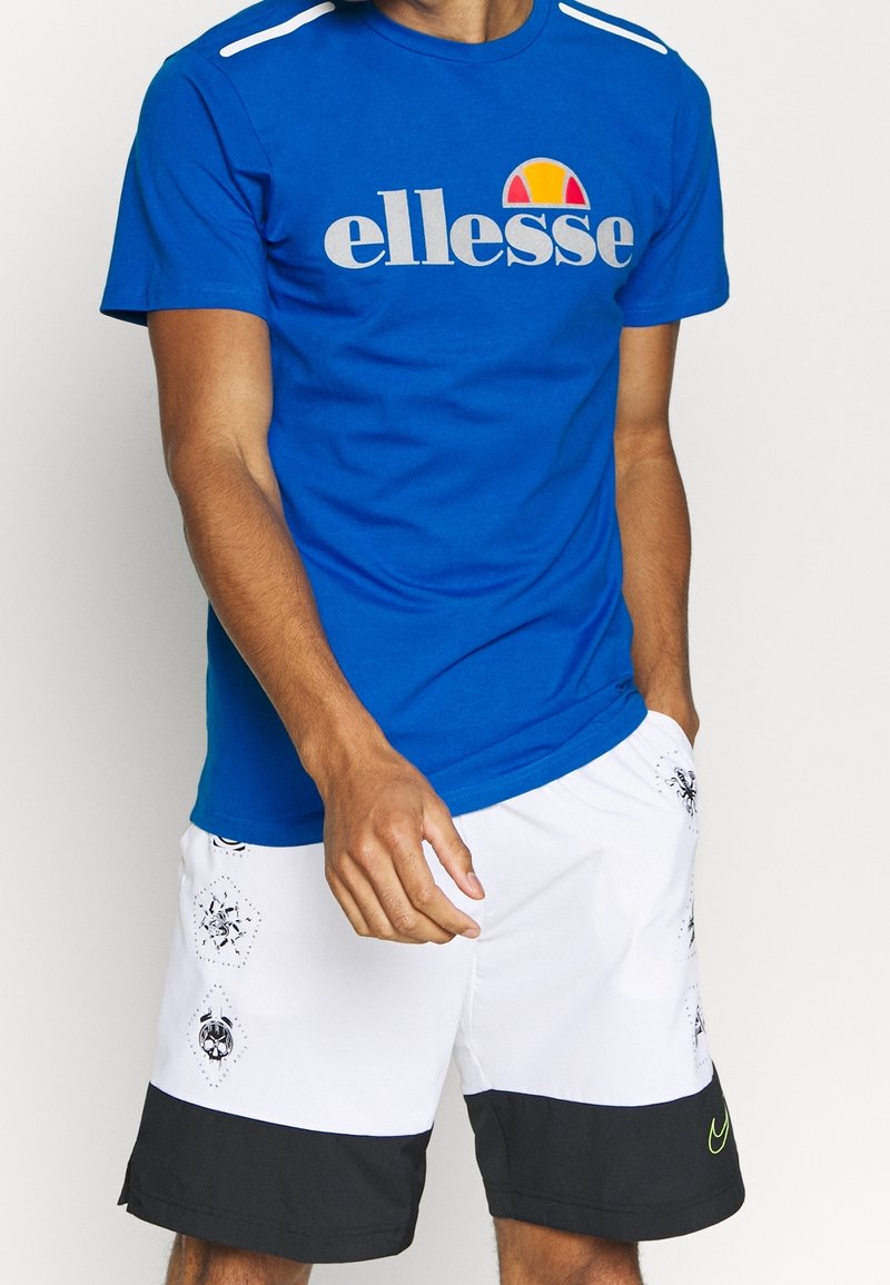 Camiseta de manga corta azul con el logo de "ellesse" en gris y naranja. Combinada con pantalones cortos blancos con dobladillo negro y acentos estampados.