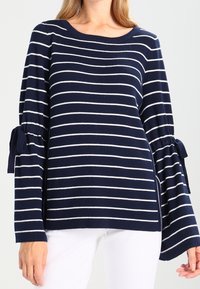 Námořnicky modrý pruhovaný svetr s bílými horizontálními pruhy, se širokými zvonovými rukávy a vázacími detaily na manžetách.