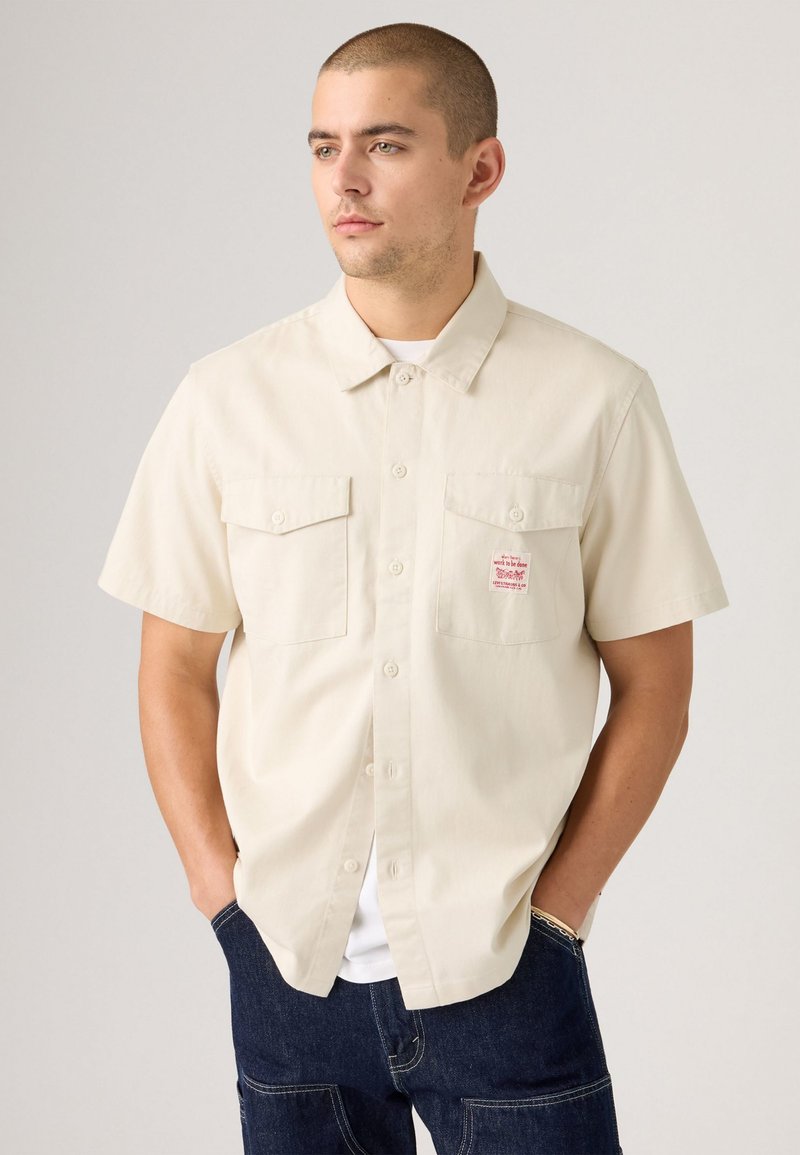 Short-sleeve beige button-up shirt with two front pockets and a small red logo patch on the left chest. Made of a smooth cotton fabric.