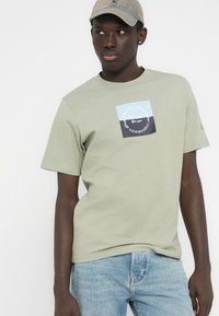 Ανοιχτό πράσινο βαμβακερό T-shirt με τετράγωνο λογότυπο σε μπλε και σκούρο γκρι, κοντά μανίκια και κλασικό στρογγυλό λαιμό.