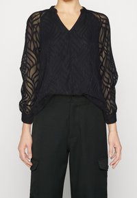 Blouse noire transparente avec un col en V et des manches longues, présentant un motif à texture ondulée et des ourlets élastiques, associée à un pantalon noir.