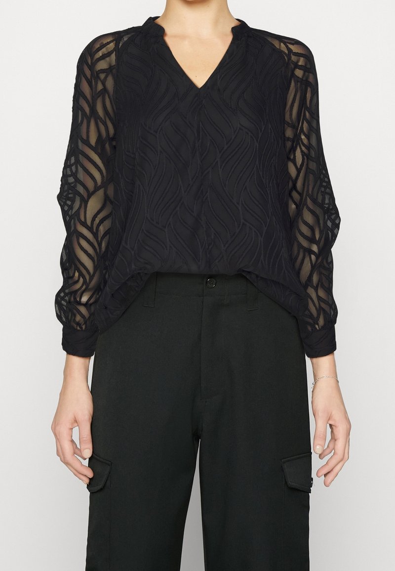 Blouse noire transparente avec un col en V et des manches longues, présentant un motif à texture ondulée et des ourlets élastiques, associée à un pantalon noir.