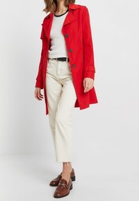 Un abrigo trench rojo con cierre de botones, gran cuello y mangas ceñidas. Combinado con unos jeans blancos y mocasines marrones de tacón bajo.