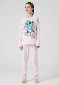 Fille aux cheveux tressés portant un pyjama rose clair avec Stitch et le texte "Just Chill", se tenant pieds nus devant un fond uni.
