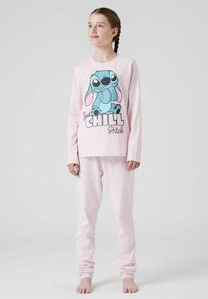 Ragazza con capelli intrecciati che indossa un pigiama rosa chiaro con Stitch e la scritta "Just Chill", in piedi a piedi nudi su uno sfondo semplice.