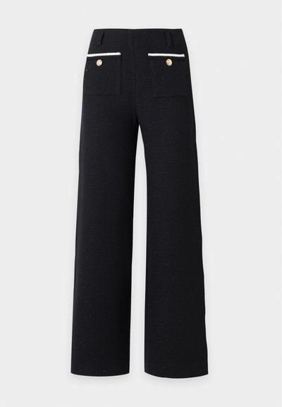 Pantalon noir à jambes larges avec poches plaquées à l’arrière, bordées de blanc et boutons dorés.