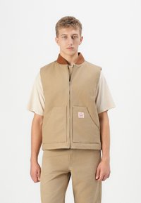 Jonge man in een beige zip-up vest over een lichtbeige shirt met korte mouwen en bijpassende beige broek, staand tegen een effen witte achtergrond.