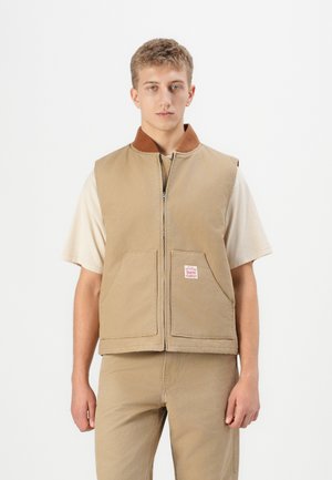Jeune homme portant un gilet zippé beige sur une chemise à manches courtes beige clair et un pantalon beige assorti, se tenant contre un fond blanc uni.