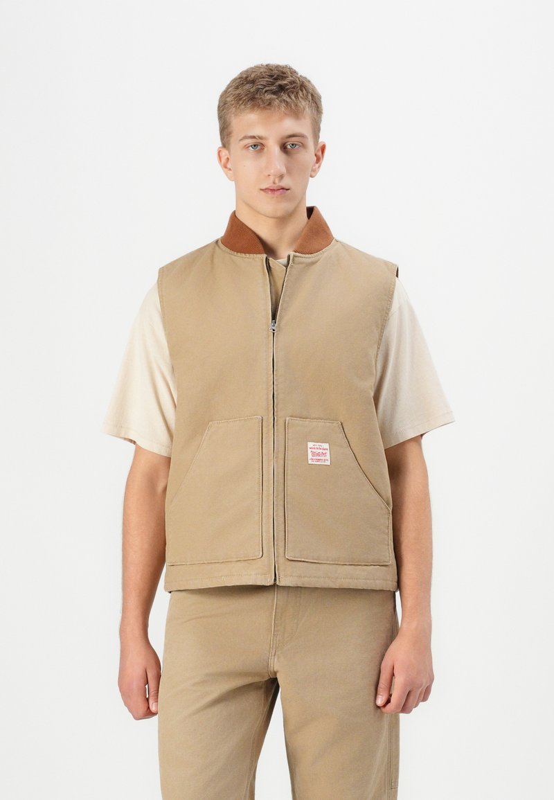 Jonge man in een beige zip-up vest over een lichtbeige shirt met korte mouwen en bijpassende beige broek, staand tegen een effen witte achtergrond.