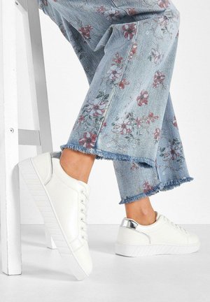 Sneakers bianchi in pelle con suola testurizzata e accenti metallici sul tallone, abbinati a pantaloni di denim chiaro a zampa d'elefante con motivi floreali.