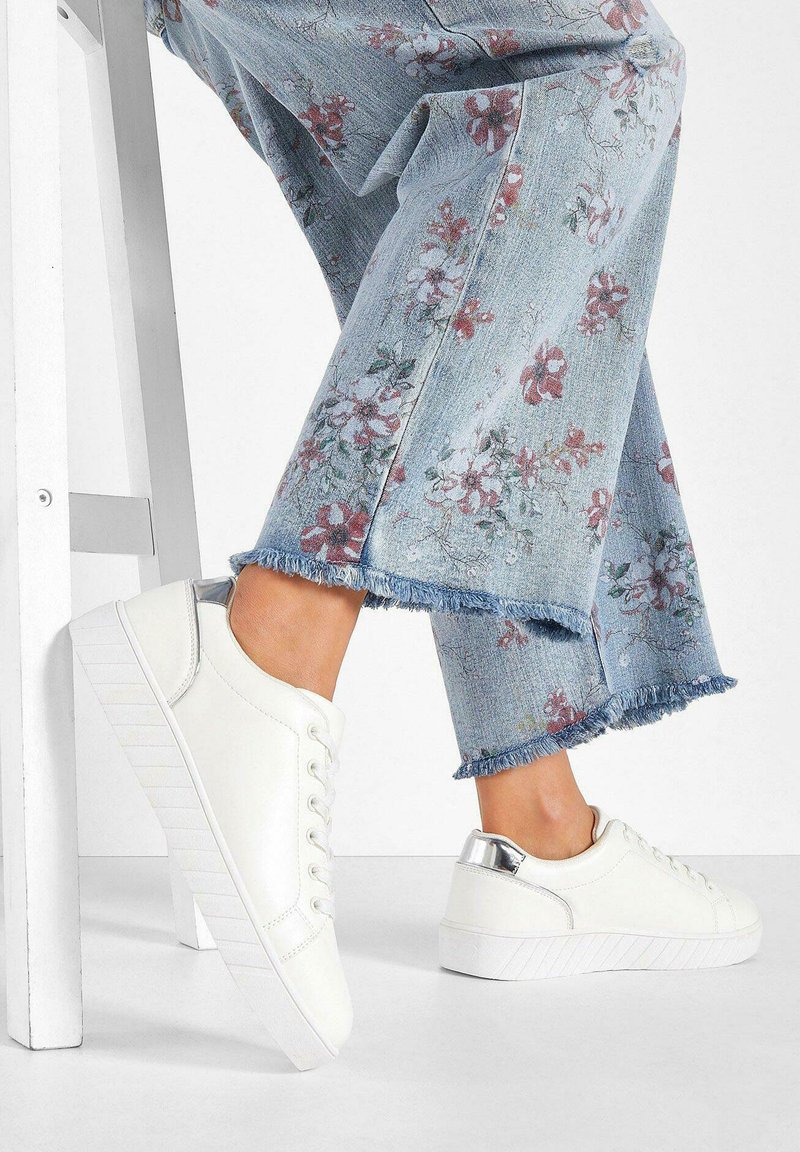 Sneakers bianchi in pelle con suola testurizzata e accenti metallici sul tallone, abbinati a pantaloni di denim chiaro a zampa d'elefante con motivi floreali.