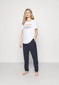 Vit kortärmad t-shirt med flerfärgad text som står "KÄRLEKENS KRAFT", tillsammans med marinblå, löst sittande byxor och barfota.