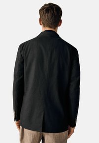 Schwarze Jacke mit tailliertem Schnitt, aus strukturiertem Stoff, mit einem Schlitz hinten und knöpfbaren Manschetten. Getragen über gestreiften Hosen.