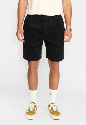 Zwarte corduroy shorts met een elastische tailleband, zijzakken en opgerolde zoom, gecombineerd met witte sokken en gele suède sneakers.