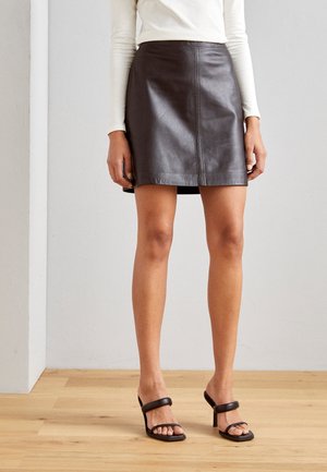 Femme portant une mini-jupe en cuir marron foncé, un haut crème à manches longues et des sandales à talons noires, debout sur un sol en bois.
