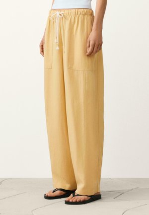 Femeie purtând pantaloni largi galbeni cu șnur în talie, buzunare frontale și sandale negre plate pe podea texturată.