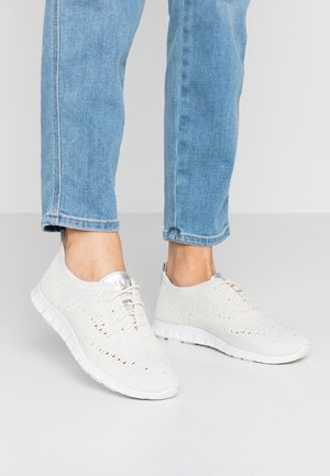Witte gebreide sneakers met veters gedragen met lichtblauwe denimjeans, getoond van de onderbenen naar beneden tegen een effen achtergrond.