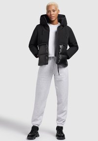 Zwarte pufferjas met capuchon; matte bovenkant en glanzende onderkant. Lichtgrijze sweatshirt eronder en grijze trainingsbroek. Zwarte sneakers.