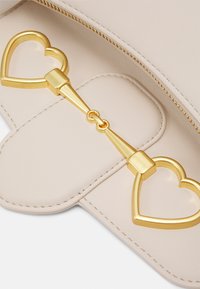 Jasnobeżowa skórzana kopertówka z złotym, sercowym zapięciem, z zamknięciem na zamek błyskawiczny, gładką fakturą i przeszyciami na klapie.