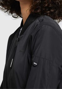 Černá bomber bunda se zipem vpředu, vroubkovaným límcem a elastickými manžetami. Na hrudi se nachází značka "Khujo" a na rukávu je nášivka.