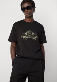 Mann iført svarte solbriller, svart T-skjorte med grønn brodert fugl og blomsterdesign, og svarte bukser, står med hendene i lommene.