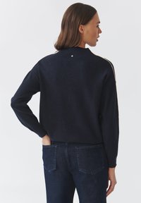 Maglione in maglia blu navy con scollo rotondo, maniche lunghe e un piccolo dettaglio decorativo sul retro, abbinato a jeans blu scuro.