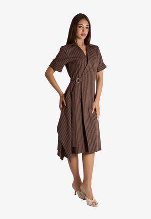 Donna che indossa un vestito avvolgente marrone a righe sottili con maniche corte e cintura, posando con una mano sui fianchi e sandali beige con tacco.