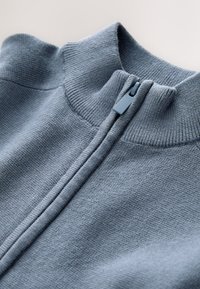 Lyseblå sweattrøje med lynlås og ribbet krave. Stoffet har en glat tekstur og indeholder en central lynlås samt sømme langs halsudskæringen.