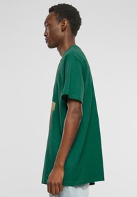 Grøn T-shirt i bomuld med afslappet pasform, korte ærmer, rund halsudskæring og et gyldent grafisk design på forsiden.