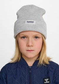 Grå beanie-hatt i ribbat material med uppvikt mudd och en central etikett. Buren med en marinblå, vadderad jacka med en logotyp-patch.
