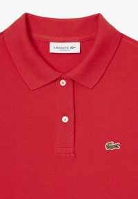 Röd polotröja i texturerad tyg, två vita knappar och en liten grön krokodil-logotyp på vänster bröst. Regular fit-design.