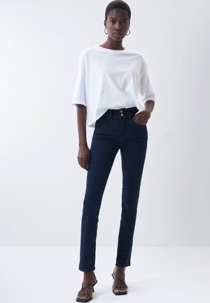 Camiseta blanca de gran tamaño, jeans ajustados azul oscuro con cierre de botón y sandalias negras de tiras, todo sobre un fondo claro.