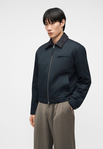 Νεαρός άνδρας με ίσια μαύρα μαλλιά, φοράει σκούρο μπουφάν με φερμουάρ και μαύρο γιακά, καθώς και μπεζ πλισέ παντελόνι.