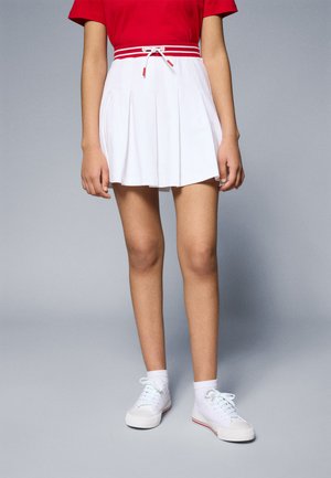 Falda blanca plisada con cintura elástica roja y rayas rojas a juego. Combinada con zapatillas blancas y calcetines hasta el tobillo. Textura de tela suave.